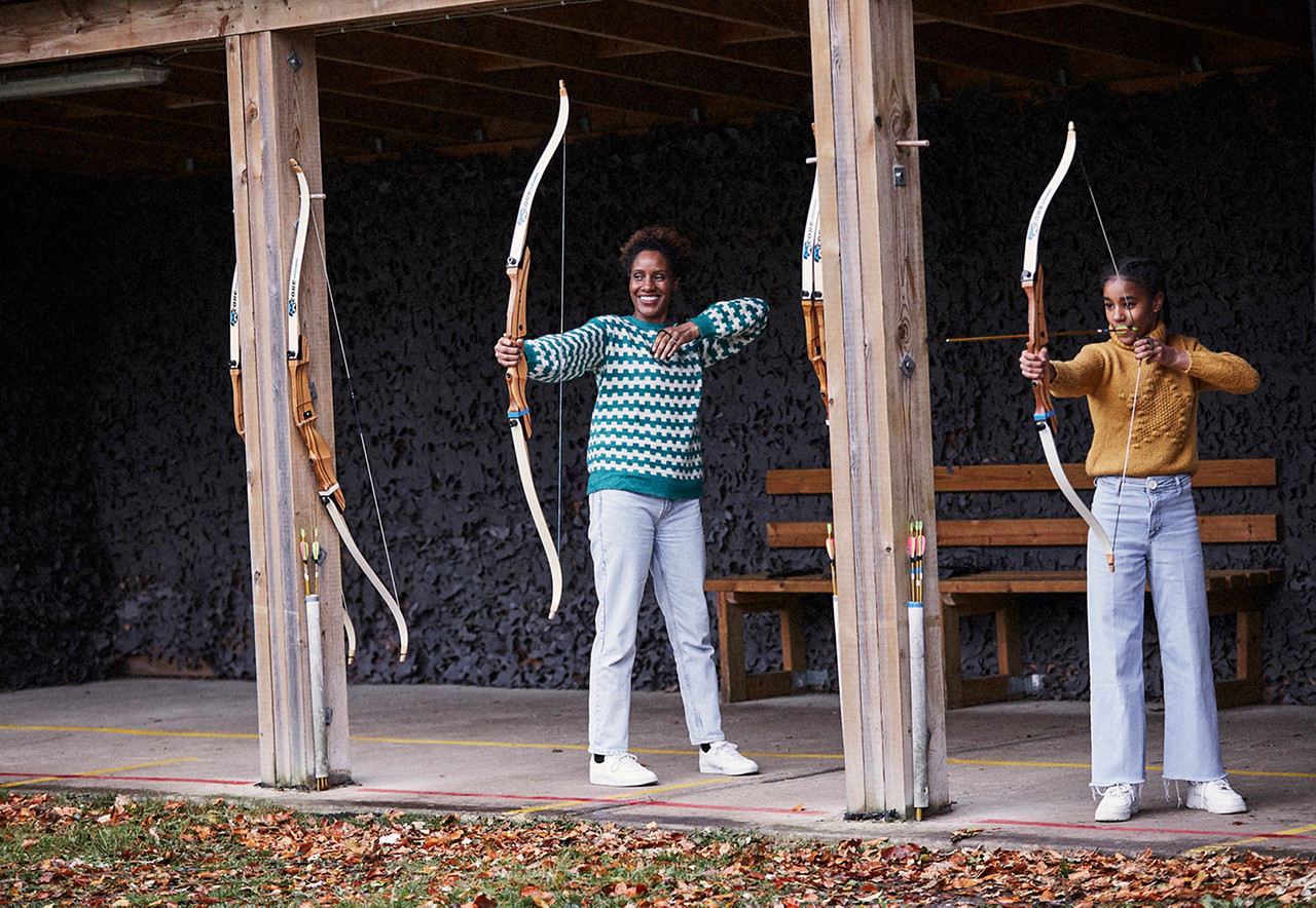Two archers draw recurve bows, one aiming while the other prepares to shoot, under a wooden covered outdoor range with benches, hanging bows, arrow quivers, and autumn leaves scattered across the concrete.