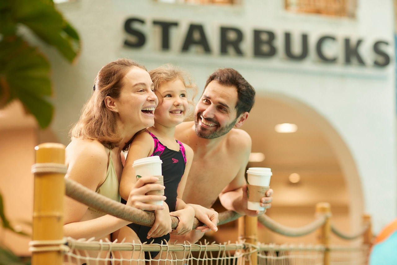 Family of three smiles and leans on a rope railing, two adults holding coffee cups, as a child in a swimsuit stands between them; indoor setting with sign reading: STARBUCKS.