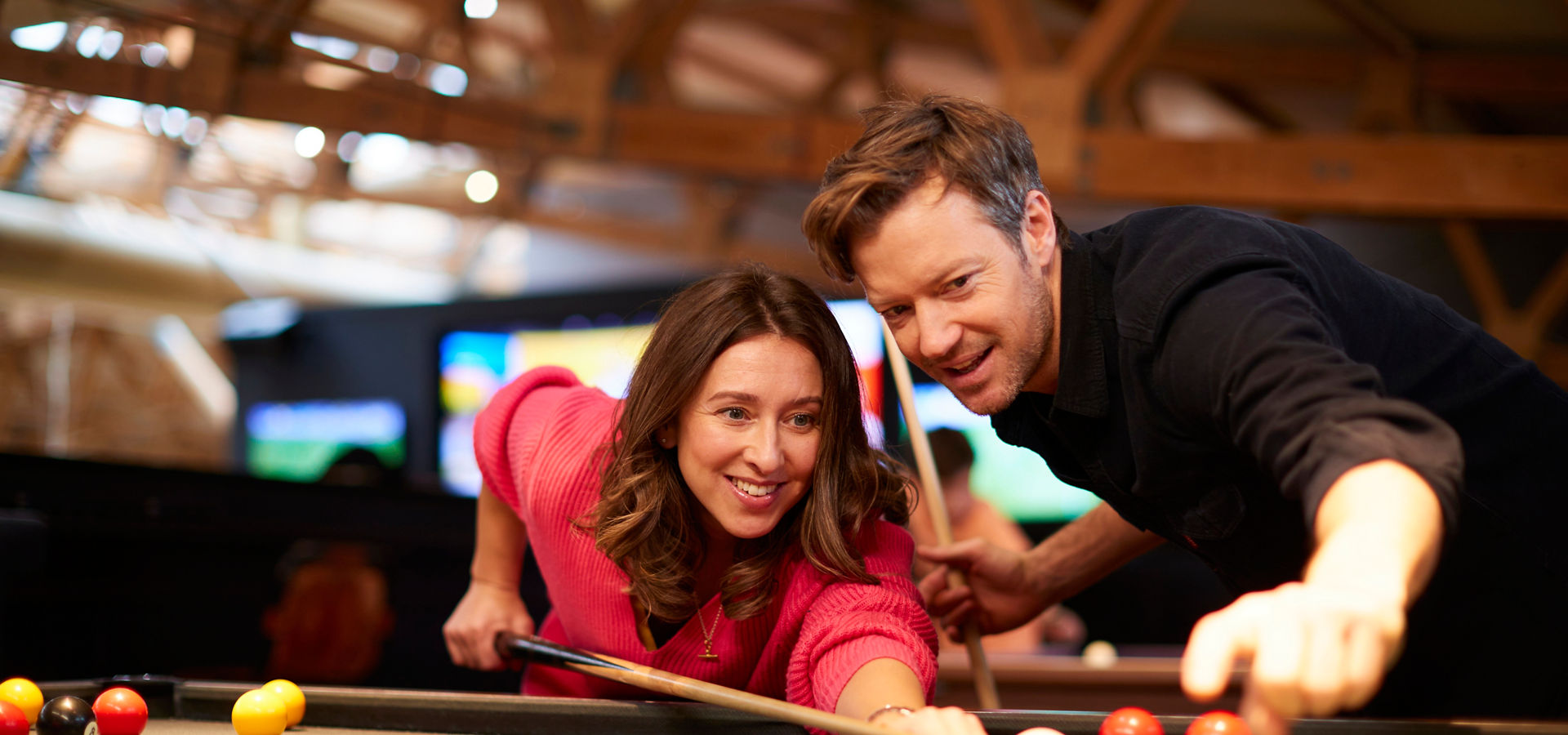 Two adults play pool; a woman aims the cue at the white ball as a man points toward the shot; colorful balls rest on felt in a warm rec hall.