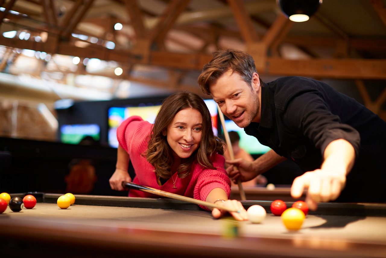 Two adults play pool; a woman aims the cue at the white ball as a man points toward the shot; colorful balls rest on felt in a warm rec hall.