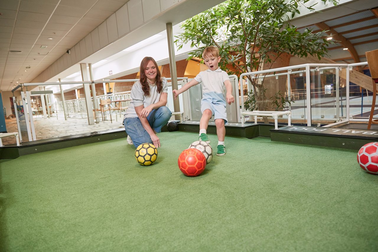 Child kicking football with mother looking on.