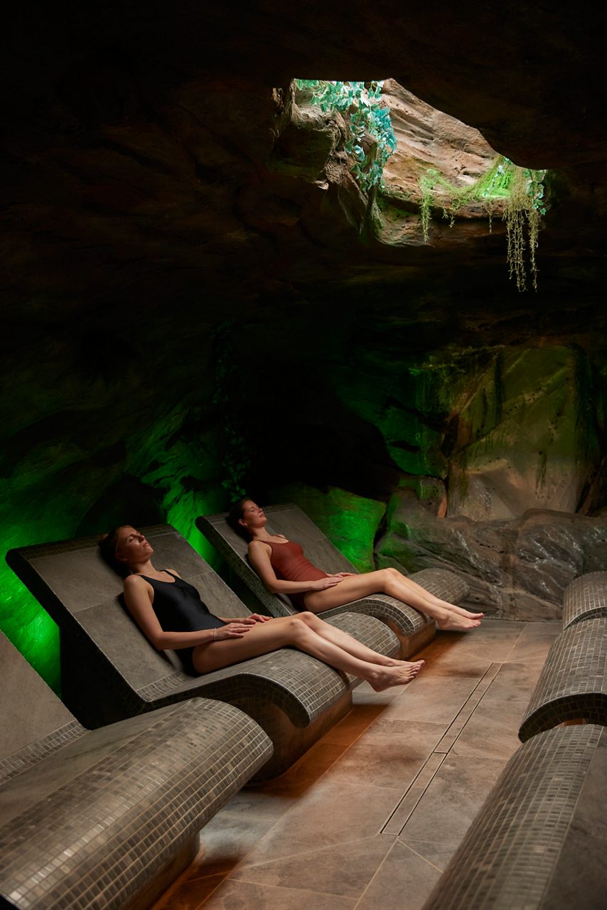 Two women laid back in the Forest Cavern room, with rock effect walls.