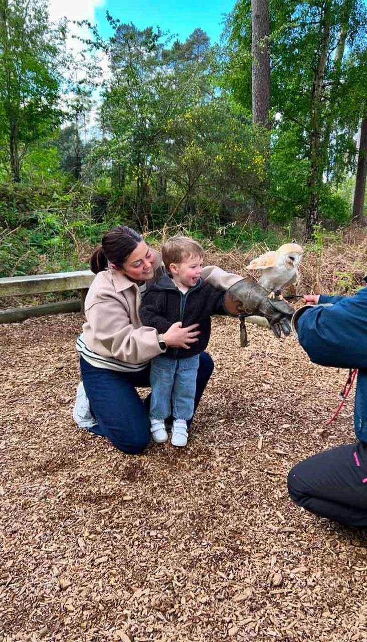 Child holds a perched owl on a leather falconry glove, smiling as an adult steadies him; instructor kneels nearby; wooded outdoor setting with woodchip ground and green trees.