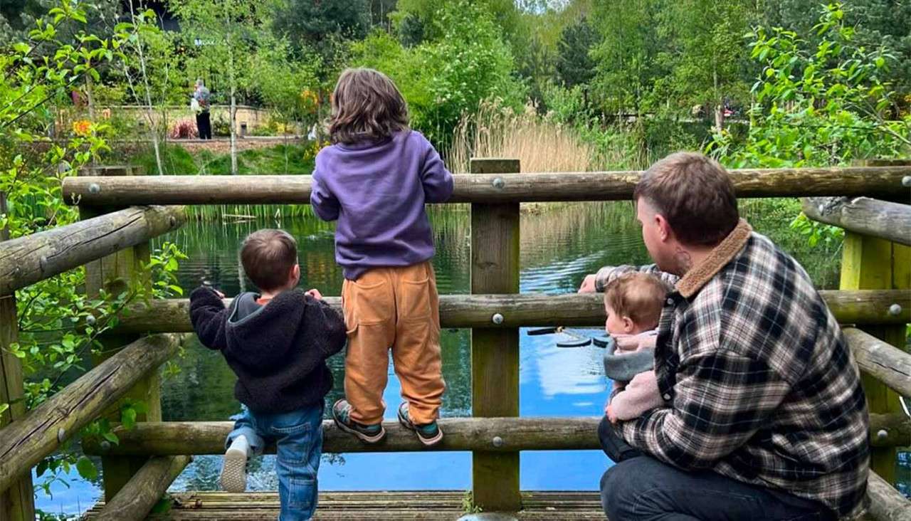 Three children and an adult sit and stand at a wooden railing, watching a calm pond with reeds, surrounded by lush trees in a park.