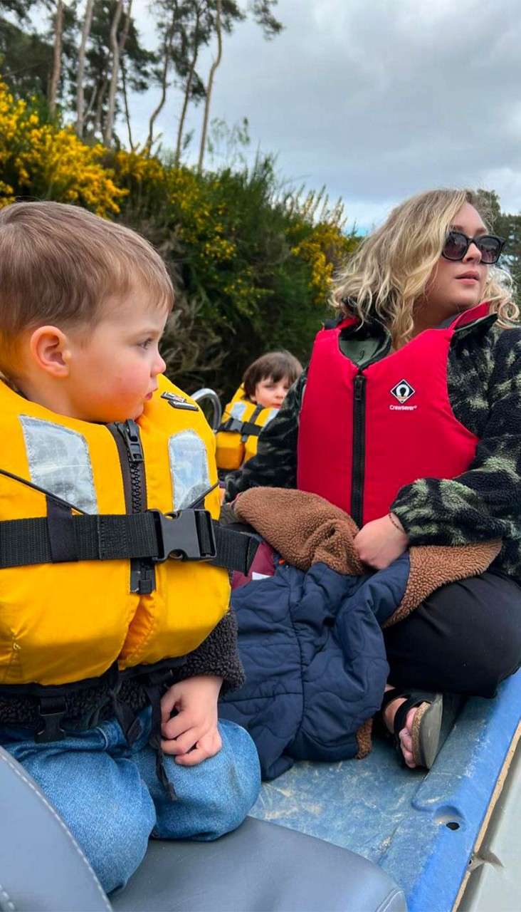 Adult and two children wear life jackets, sit and look sideways while riding in a small open boat; one holds blankets. Bushy shoreline and cloudy sky behind. Text: Crewsaver.