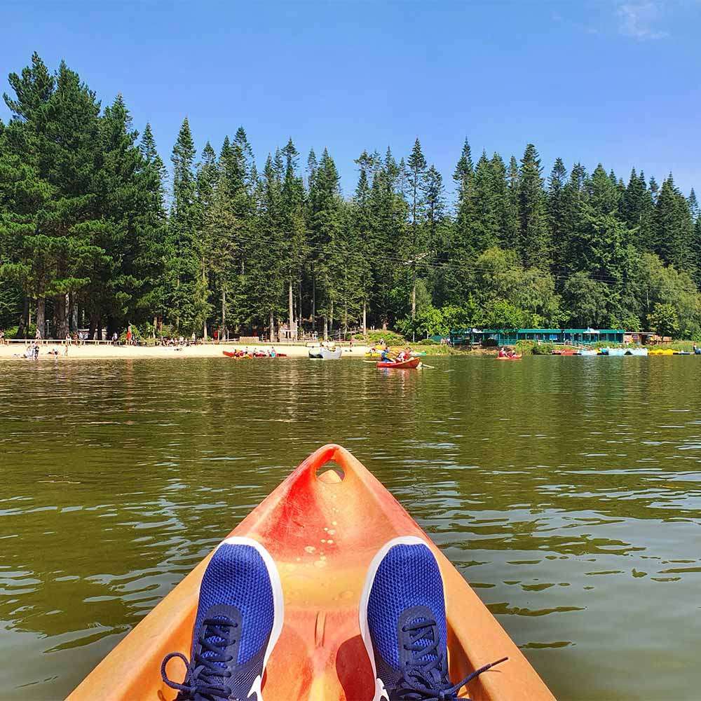 Kayak bow with sneakered feet points forward, gliding on calm lake water; surrounding context includes other kayaks and a sandy shore lined with tall evergreen trees beneath a clear blue sky.