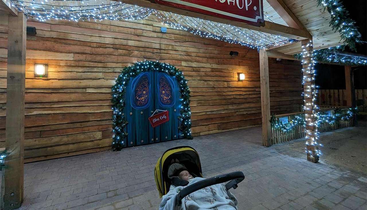 Blue arched door glows with Christmas lights, framed by garland, on a wooden building; a baby sleeps in a stroller in front. Text: “Elves Only” and “SHOP.”