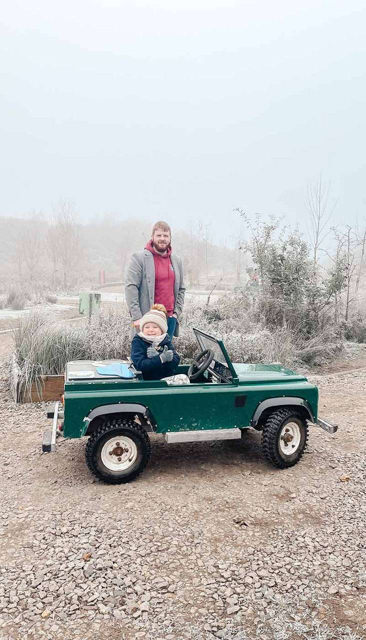 Child sits in a small green toy jeep, smiling and giving thumbs-up; adult stands behind watching. Context: foggy, frosty outdoor park with gravel path, sparse winter vegetation and muted hills.