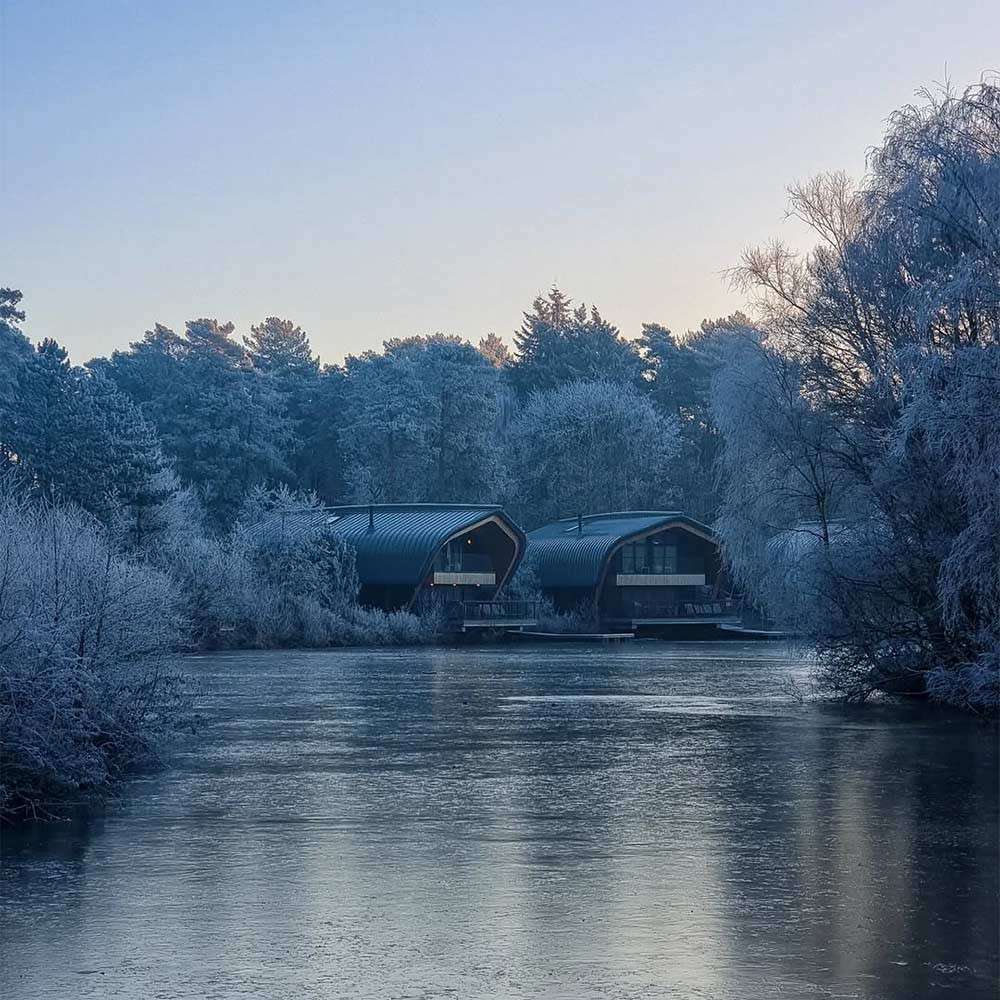 Two modern cabins sit beside a frozen lake, reflecting faintly, surrounded by frost-covered trees at dawn under a clear, pale sky.