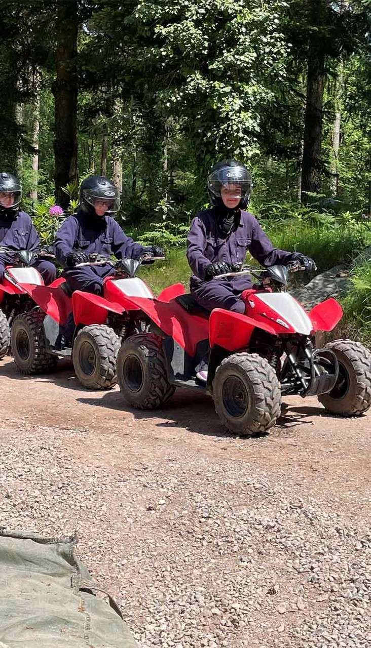 ATV riders drive in a line along a gravel path, wearing dark clothing and helmets, through a sunlit forest with dense green foliage.