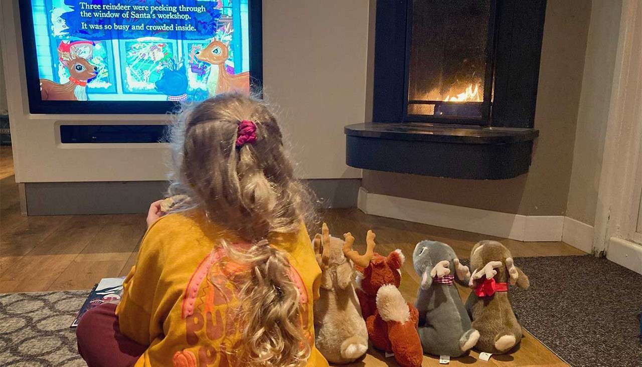 Child watches a TV Christmas cartoon; stuffed animals sit on the floor; fireplace glows in a cozy living room. On-screen text: "Three reindeer were peeking through the window of Santa's workshop. It was so busy and crowded inside."
