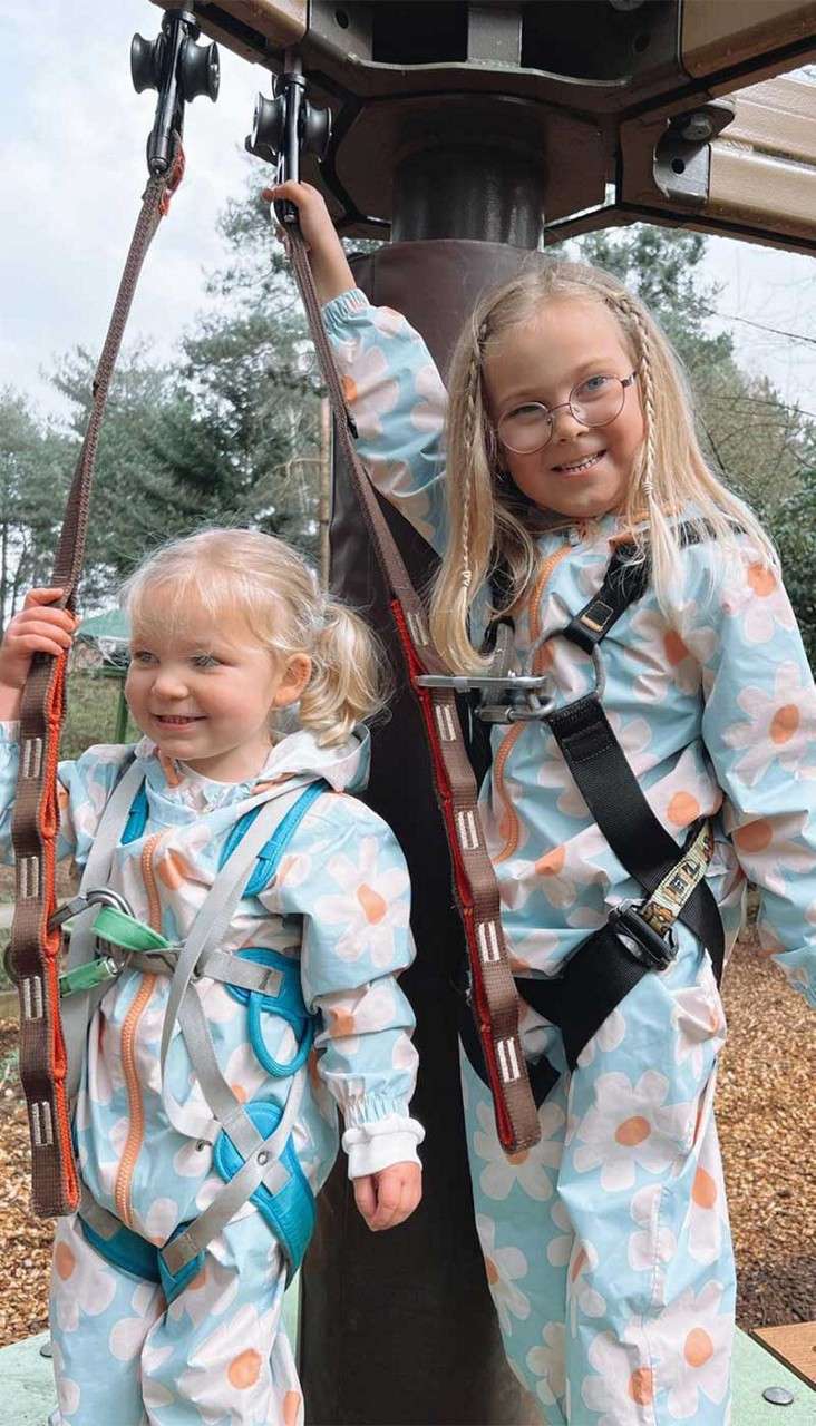 Two children smile, gripping safety straps and wearing matching floral jumpsuits and harnesses. They stand beneath an outdoor rope-course apparatus by a central pole, with trees and wood-chip ground behind.