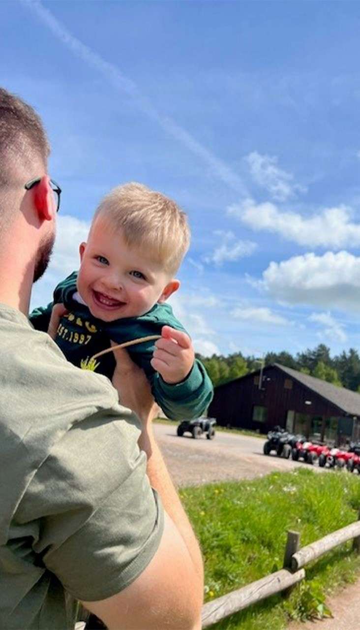 Smiling toddler faces the camera while held aloft by an adult, gripping a straw. Behind them: rustic building, parked ATVs, wooden fence, and grassy field beneath a bright blue sky.