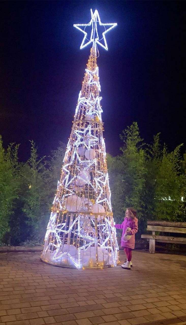 Illuminated cone-shaped Christmas tree sculpture glows white, topped with a star. A child reaches toward it in curiosity, standing on brick pavement in a nighttime park beside benches and shrubs.