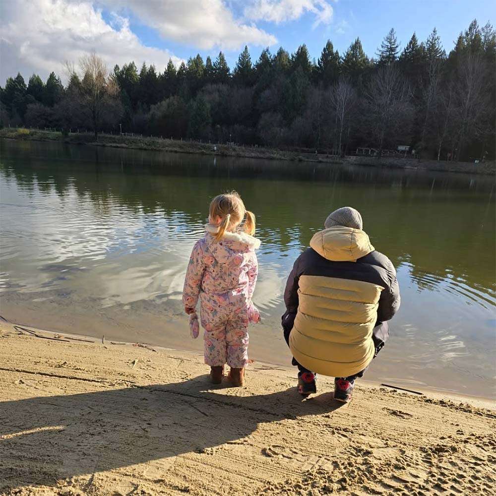 Two bundled figures—one child standing, one adult crouching—watch ripples on a calm lake, on a sandy shore, with evergreen forest backdrop and bright winter sunlight casting long shadows.