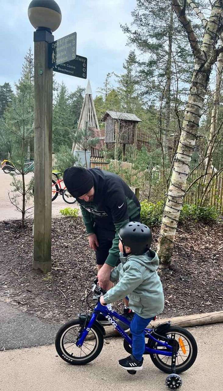 Child on blue bike with training wheels pedals as adult steadies him; wooded park path beside a signpost reading: Cycle & Footway, Footway, Cycle Centre, Beechwood.