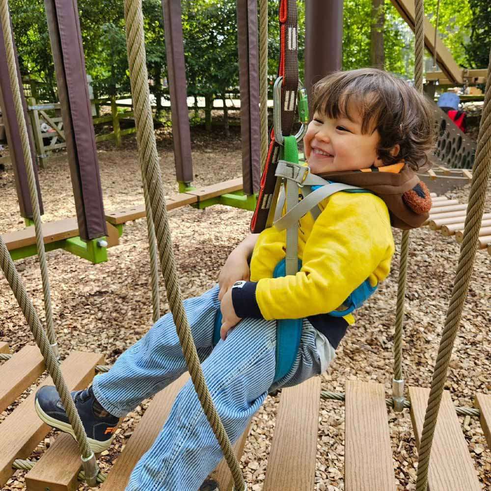 Child smiles while sitting in a safety harness, clipped to ropes, gliding over wooden planks. Context: outdoor adventure playground with rope bridges, mulch ground, green trees and equipment in background.