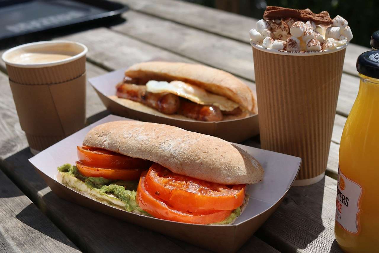 Two ciabatta sandwiches rest in cardboard trays—front with tomato and avocado, back with sausage and fried egg—accompanied by coffee, orange juice labeled “ORANGE,” and marshmallow-topped hot chocolate on a wooden picnic table.