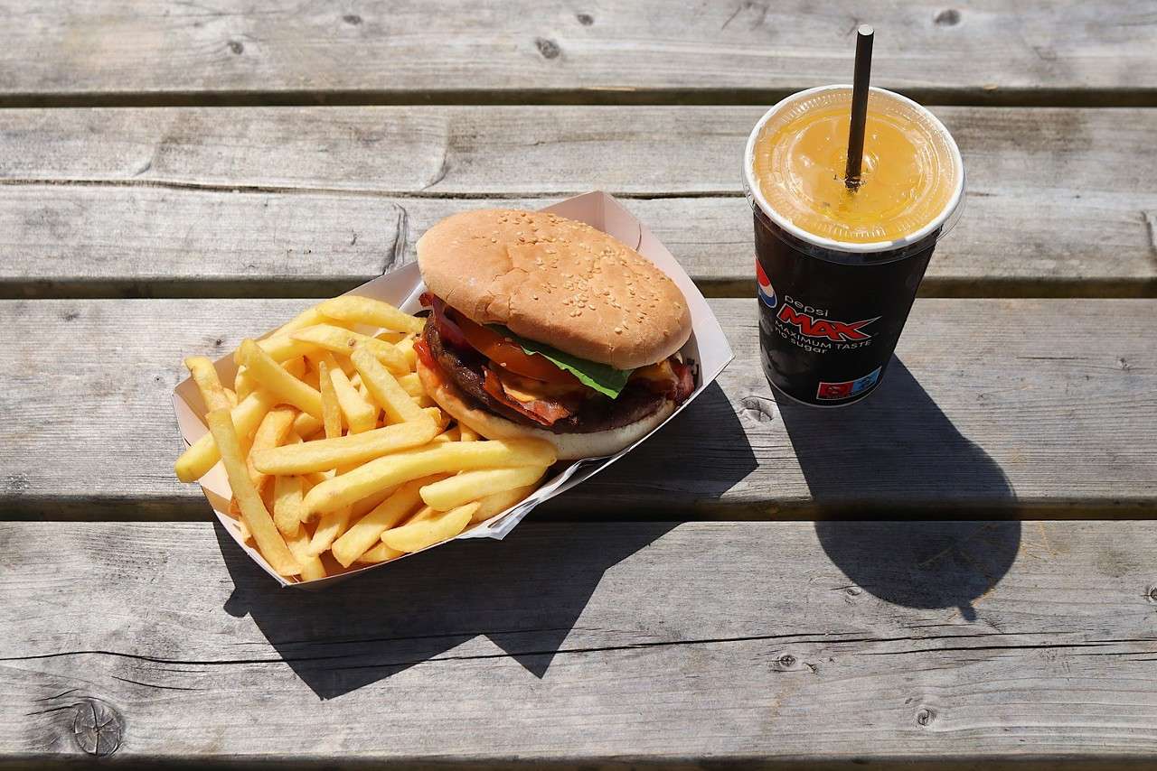 Burger with lettuce, cheese, and bacon sits in a paper tray beside French fries; a lidded drink with straw stands nearby on a sunlit wooden picnic table. Text: "pepsi MAX maximum taste".