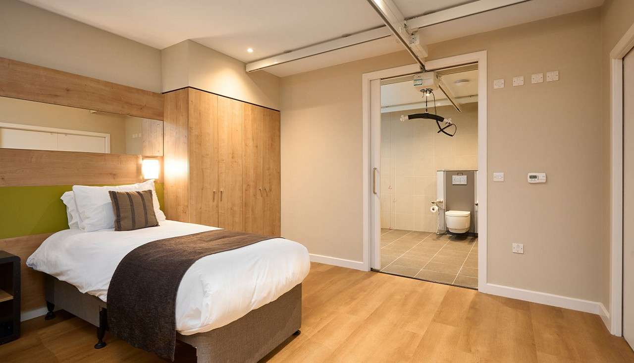 Single bed with white linens rests beside wooden wardrobes; overhead hoist rail runs into an open doorway; beyond, accessible bathroom shows a wall-mounted toilet. Warm lighting and wood floors throughout.