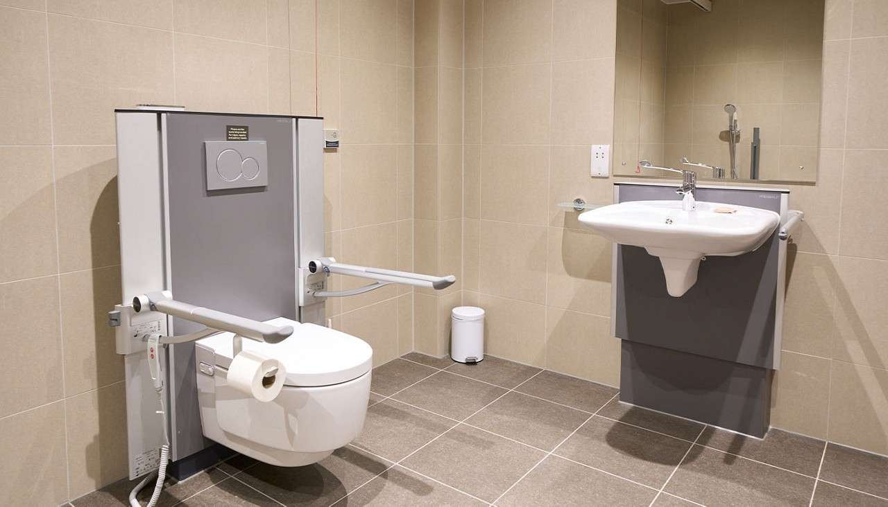 Accessible toilet features fold-down support rails and bidet controls; a wall-mounted sink with mirror sits nearby; small trash bin and tiled, spacious bathroom context designed for wheelchair access.