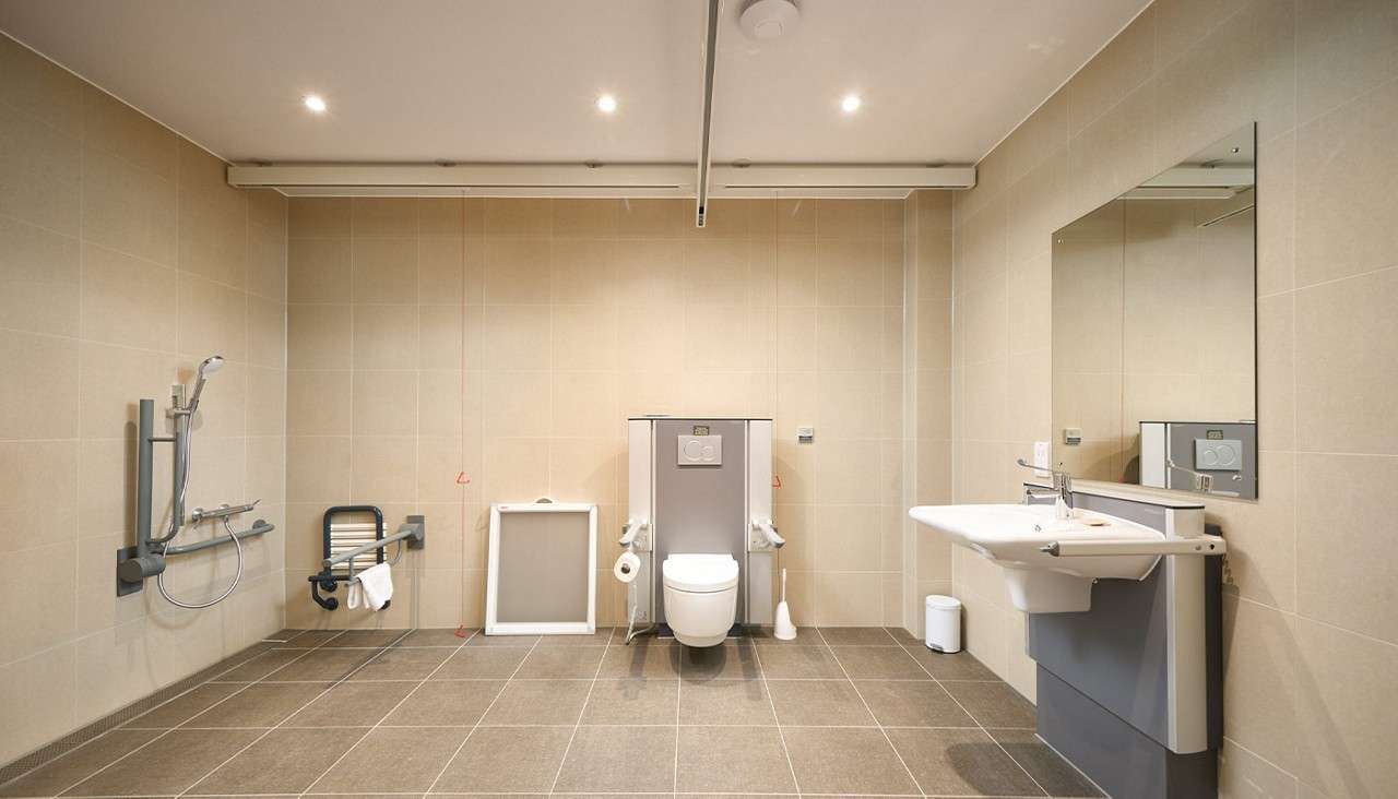 Wheelchair-accessible toilet anchors the room, flanked by support rails; nearby, a roll-in shower and wall-mounted sink sit within a tiled bathroom featuring grab bars, emergency pull cords, and ceiling hoist.