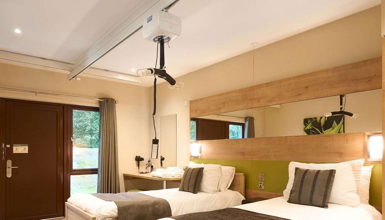 Ceiling-mounted patient lift hangs from a track, positioned over two neatly made twin beds; in a modern accessible room with wood headboard mirror, green accent wall, desk, window, and lighting.