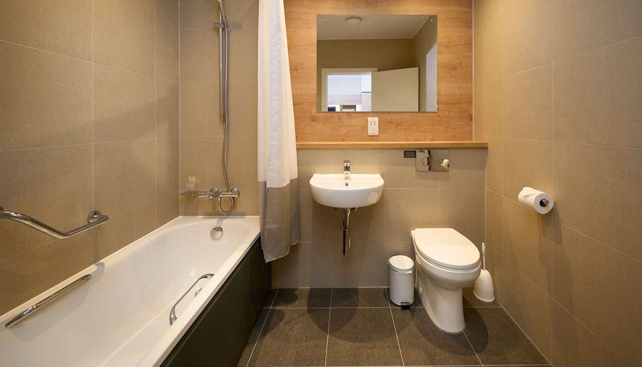 Bathtub with shower curtain partially drawn, adjacent to a wall-mounted sink and toilet, in a tiled bathroom featuring a mirror, wooden backsplash, grab bars, trash can, and toilet paper holder.