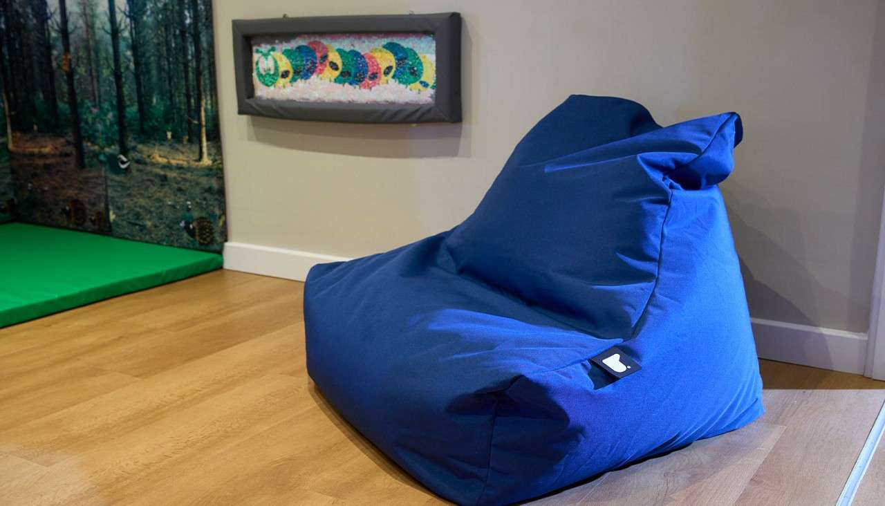 Blue beanbag chair rests on a wooden floor, angled toward a corner. Nearby, a green floor mat and forest-themed wall panel sit beneath a framed, colorful fabric artwork.