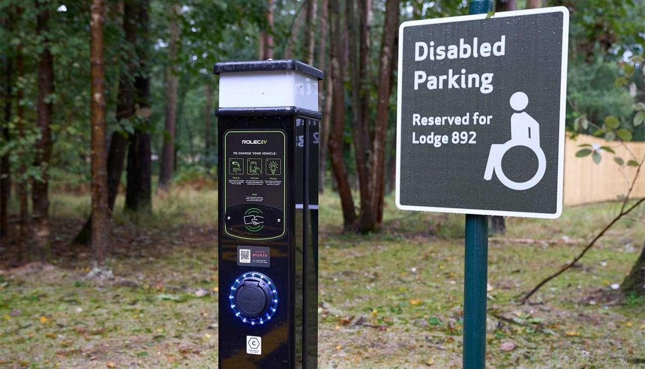 Electric vehicle charger stands beside a parking sign in a wooded area. Text: “Disabled Parking Reserved for Lodge 892.” Charger text: “ROLEC EV,” “TO CHARGE YOUR VEHICLE,” “RFID,” “MONTA,” “ROLEC,” and “C.”