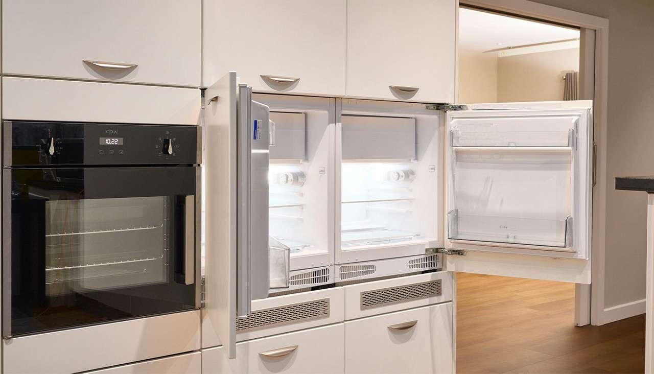 Open built-in refrigerator and freezer reveal empty, lit shelves; context: modern white kitchen cabinetry with adjacent black oven labeled CDA, its display reading 10:22, set above drawers and hardwood floor.