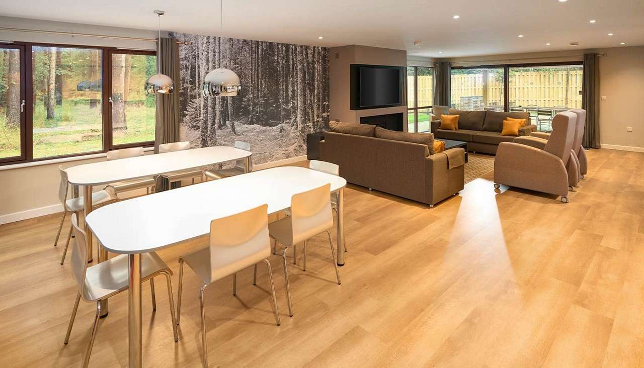 Open-plan lounge-diner features white tables with chairs and brown sofas arranged around a TV; pendant lights hang above; large windows overlook a fenced garden and forest mural decorates one wall.