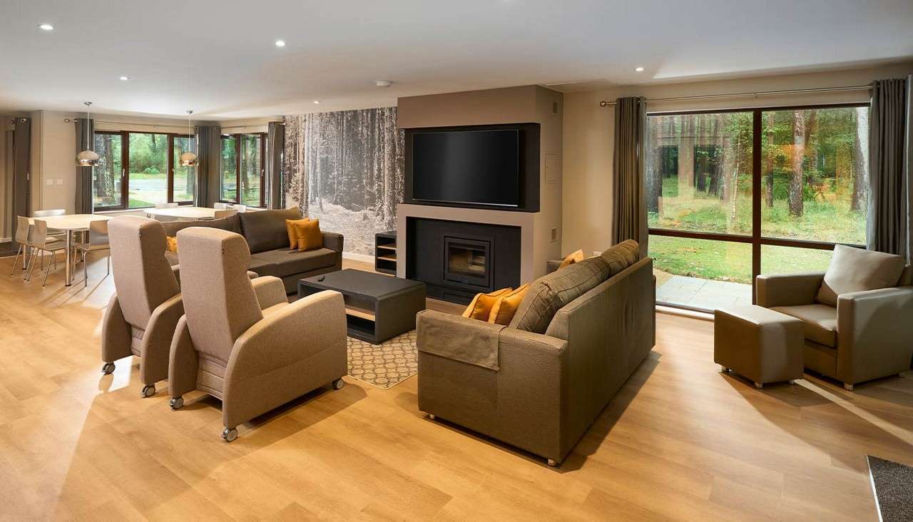 Living room seating clusters arranged around a fireplace and wall-mounted TV, overlooking wooded scenery through large windows; adjacent dining area and pendant lights, neutral tones on wooden floors.