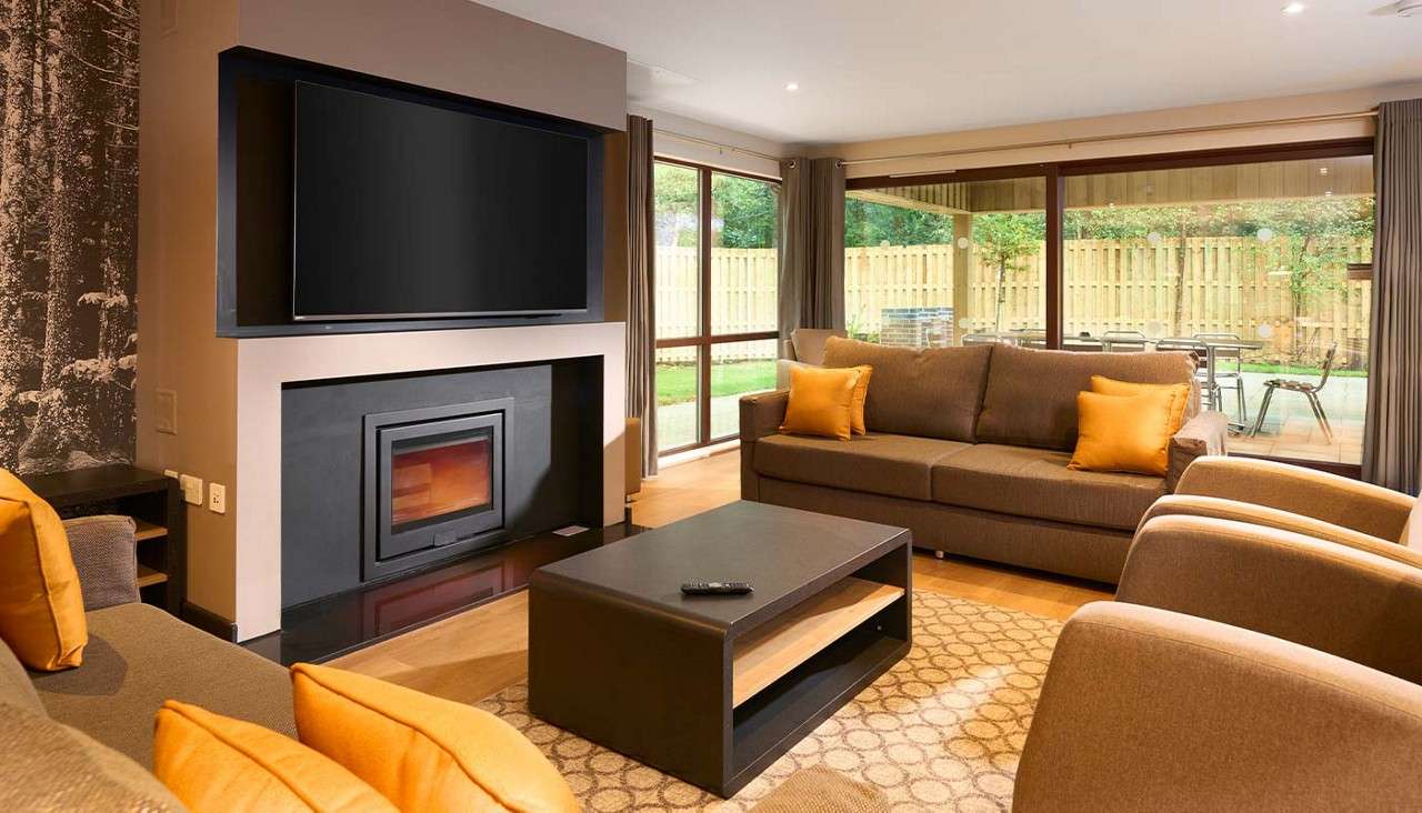 Brown sofas with orange cushions face a wall-mounted TV above a fireplace, arranged around a low coffee table; floor-to-ceiling windows reveal a fenced patio and green yard.