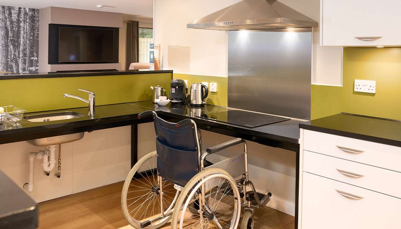 Wheelchair rolls beneath a lowered countertop, positioned at an induction cooktop and sink. Surrounding kitchen features lime-green backsplash, stainless range hood, kettle and coffee maker, open knee space, and white cabinets.