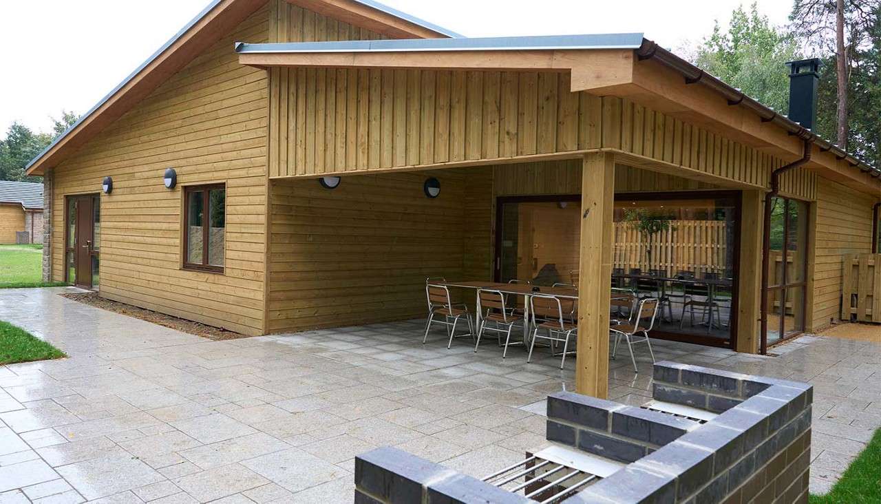Wooden cabin shelters metal tables and chairs under a covered patio. Context: paved courtyard, built-in brick barbecue in the foreground, large windows and doors, grassy edges, and trees beyond.