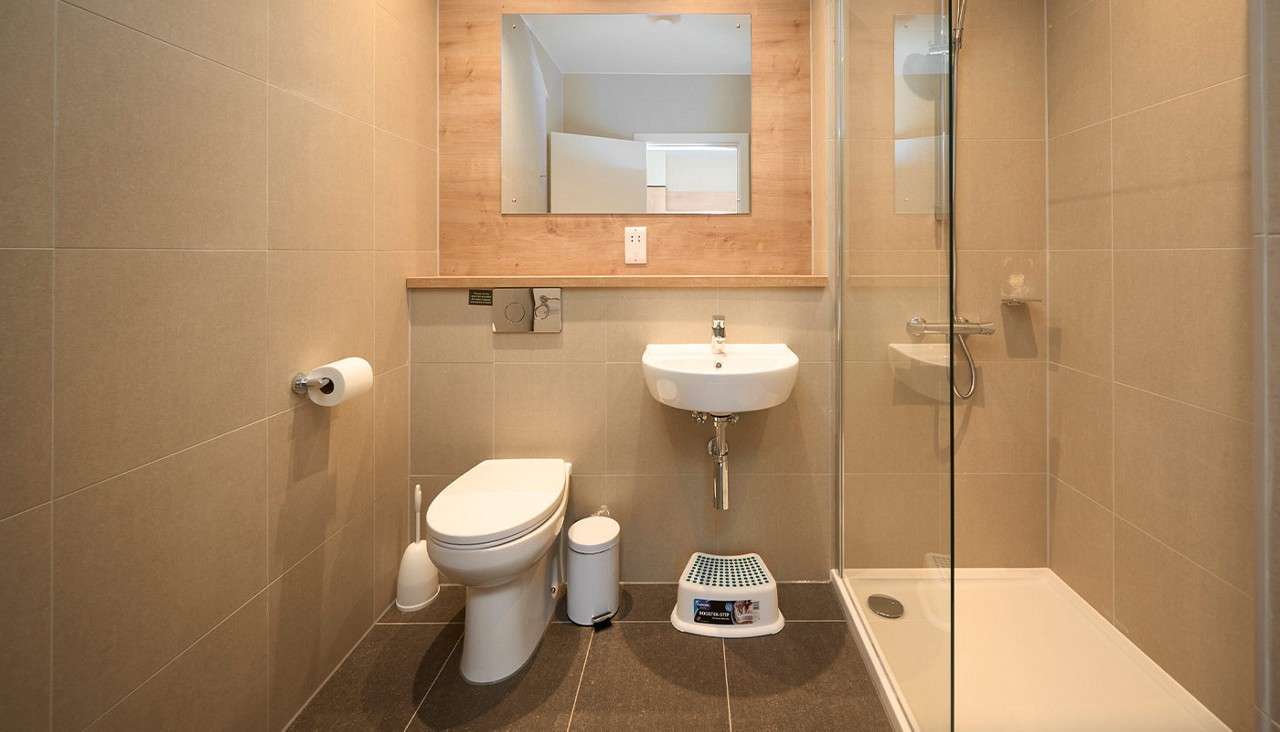 Toilet and wall-mounted sink occupy the center, flanked by a glass shower. Beige-tiled bathroom includes mirror, wooden shelf, toilet roll, brush, bin, and a small step stool below the sink.