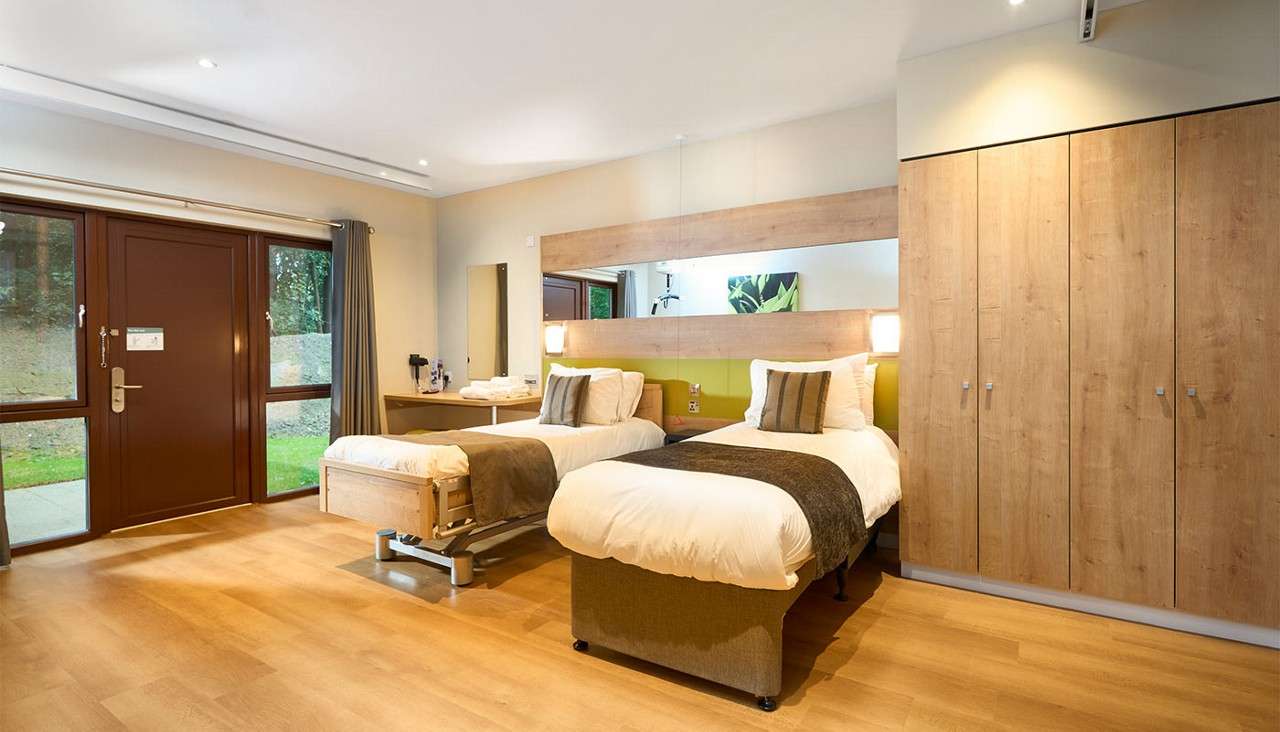 Twin beds sit side-by-side, neatly made with white linens and brown runners, in a modern room featuring wooden floor, large mirror, built-in wardrobes, desk with kettle, and door/windows opening to greenery.