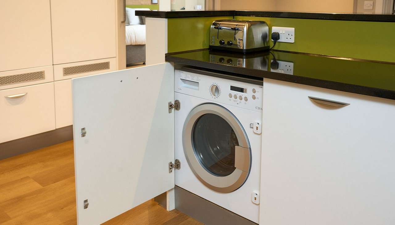 Front-loading washing machine sits built into a kitchen cabinet, revealed by an open white door; countertop above holds a chrome toaster near a wall outlet, with green backsplash. Text: CDA.