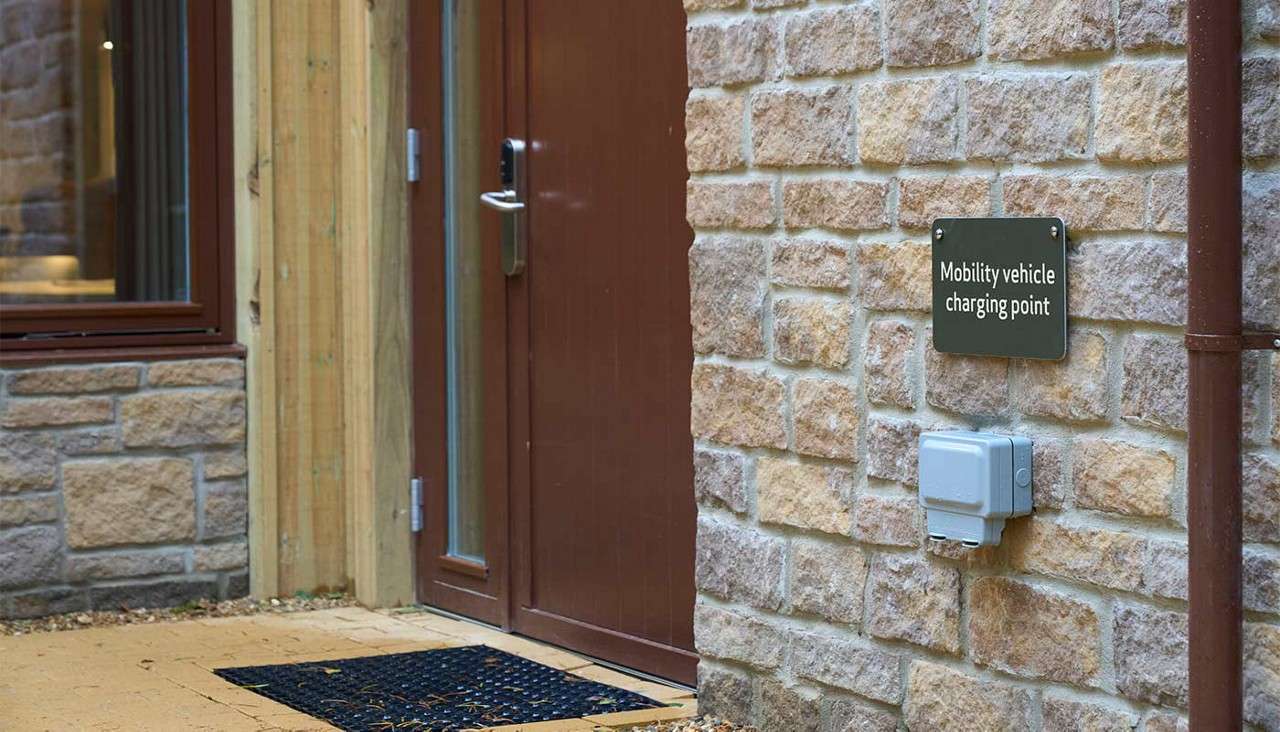 Wall-mounted charging socket indicates “Mobility vehicle charging point,” positioned beside a brown door on a stone building exterior with gutter pipe and doormat.