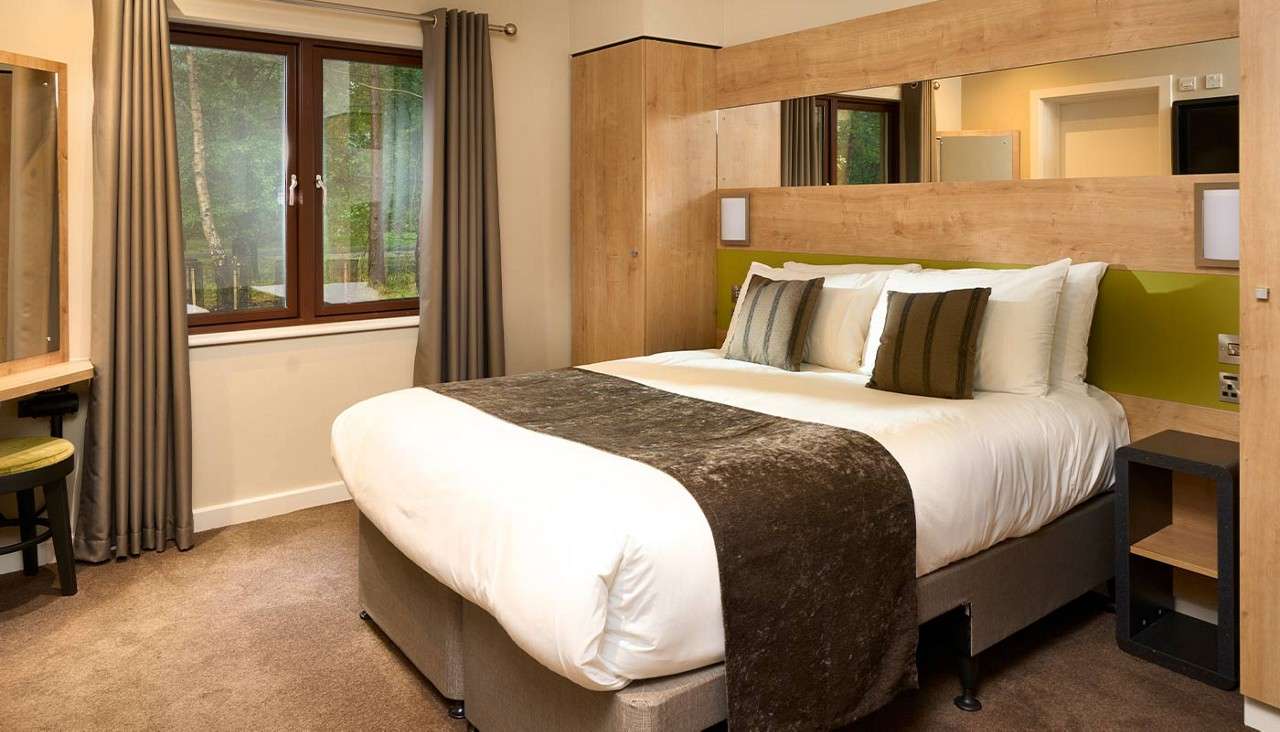 A neatly made double bed anchors the room, topped with pillows and a brown throw, beside wooden wardrobes and windows with gray curtains, overlooking trees; desk and nightstand nearby.