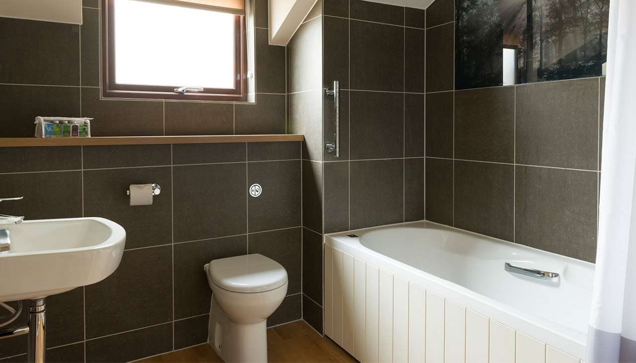 Bathtub anchors a compact bathroom, resting beside dark-tiled walls; nearby a toilet and sink face a wood-framed window, with a shelf of toiletries and a shower curtain framing the tub.