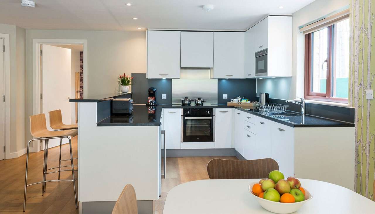Modern kitchen with white cabinets and black countertops; centered oven; toaster and coffee maker on island; dish rack by sink; foreground dining table with fruit bowl; stools and window.