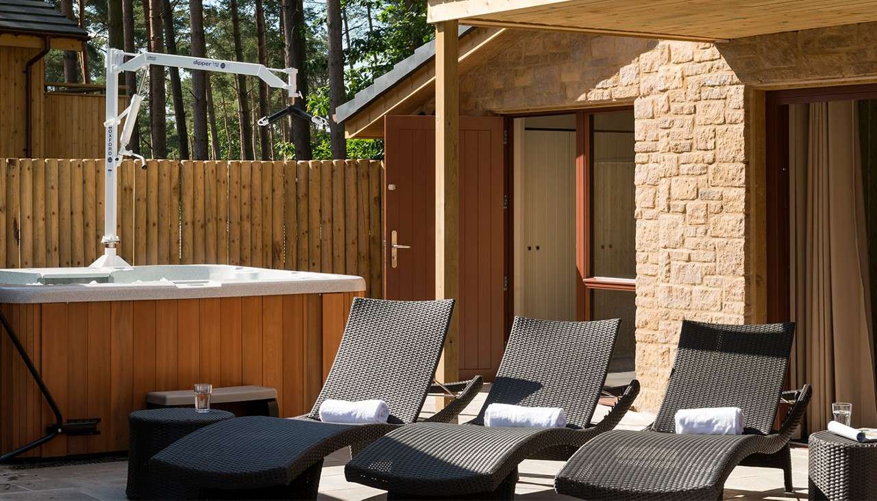 Hot tub hoist stands beside a spa on a patio with four wicker loungers holding towels; glasses nearby. Wooden fence and stone lodge in forested setting. Text: “dipper”, “OXFORD”.