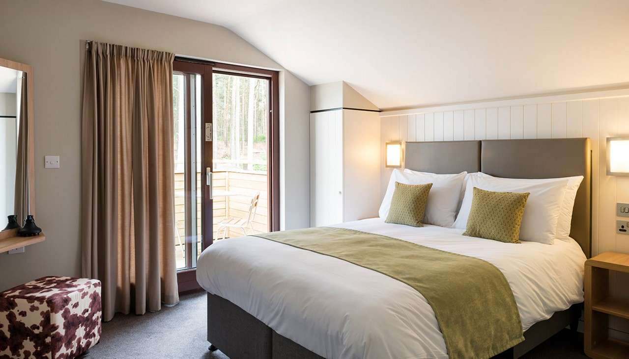 Bed with white linens and green throw sits neatly made, flanked by glowing wall lamps; sliding glass door reveals patio chairs and forest; wardrobe, curtain, and ottoman complete the room.