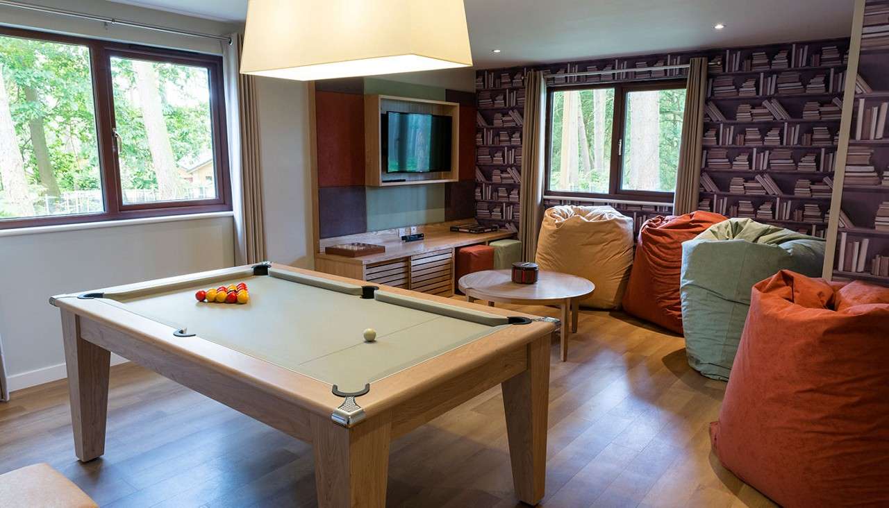 Pool table sits ready, balls racked and cue ball set, in a cozy game room with beanbag chairs, round coffee table, wall-mounted TV, pendant light, wood floors, and forest-view windows.