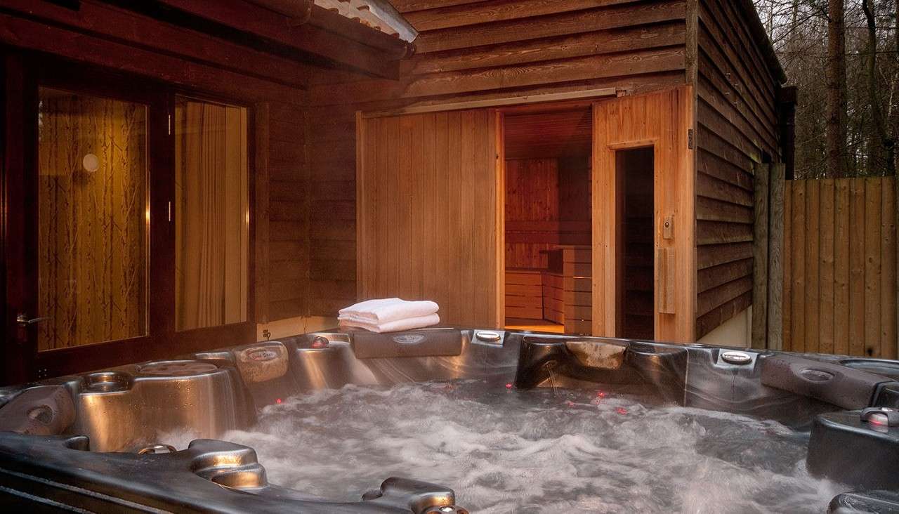 Outdoor hot tub bubbling; steam rises and folded towels rest on the edge; beside it, a wooden cabin with an open sauna door glows warmly, enclosed by a fence and trees.
