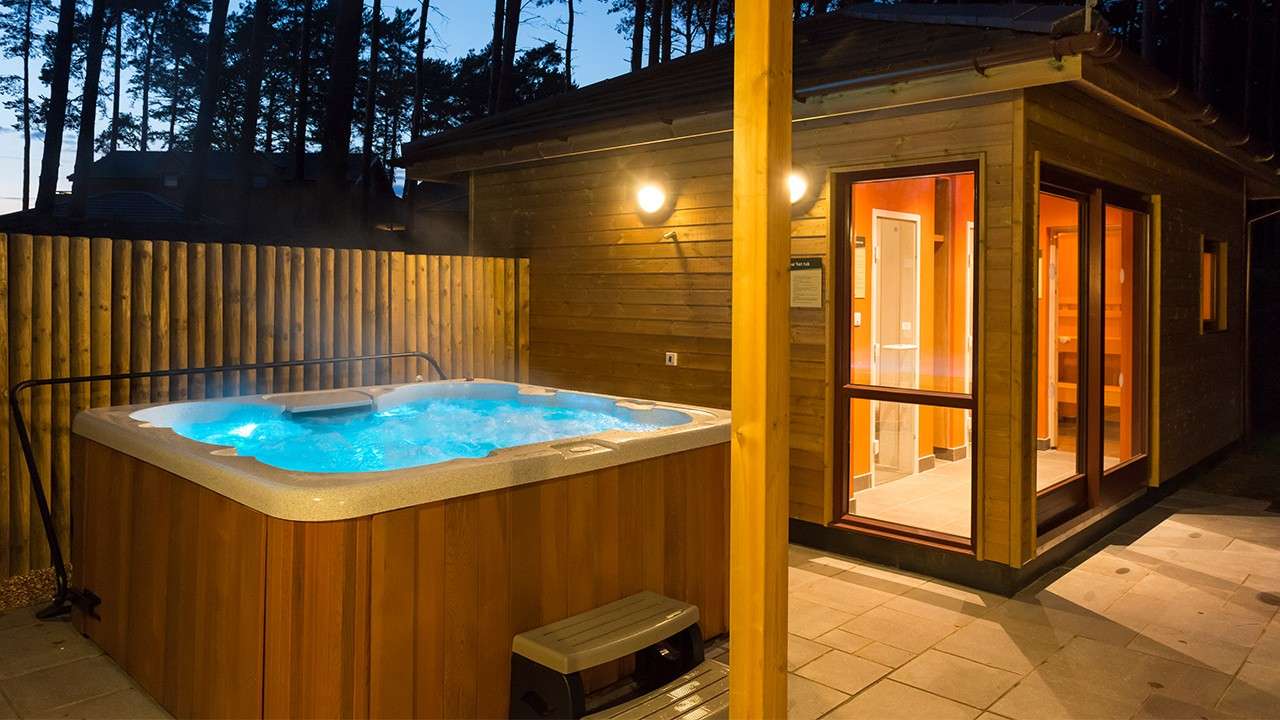 Hot tub bubbles and steams, illuminated blue, beside a lit wooden cabin. A step stool sits nearby. Wooden fence encloses a patio, with tall pine trees silhouetted at dusk.
