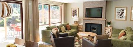 Green sofas and chairs circle a round wooden table, facing a fireplace with wall-mounted TV; large windows and a sliding door open to a patio, amid wood accents and framed art.