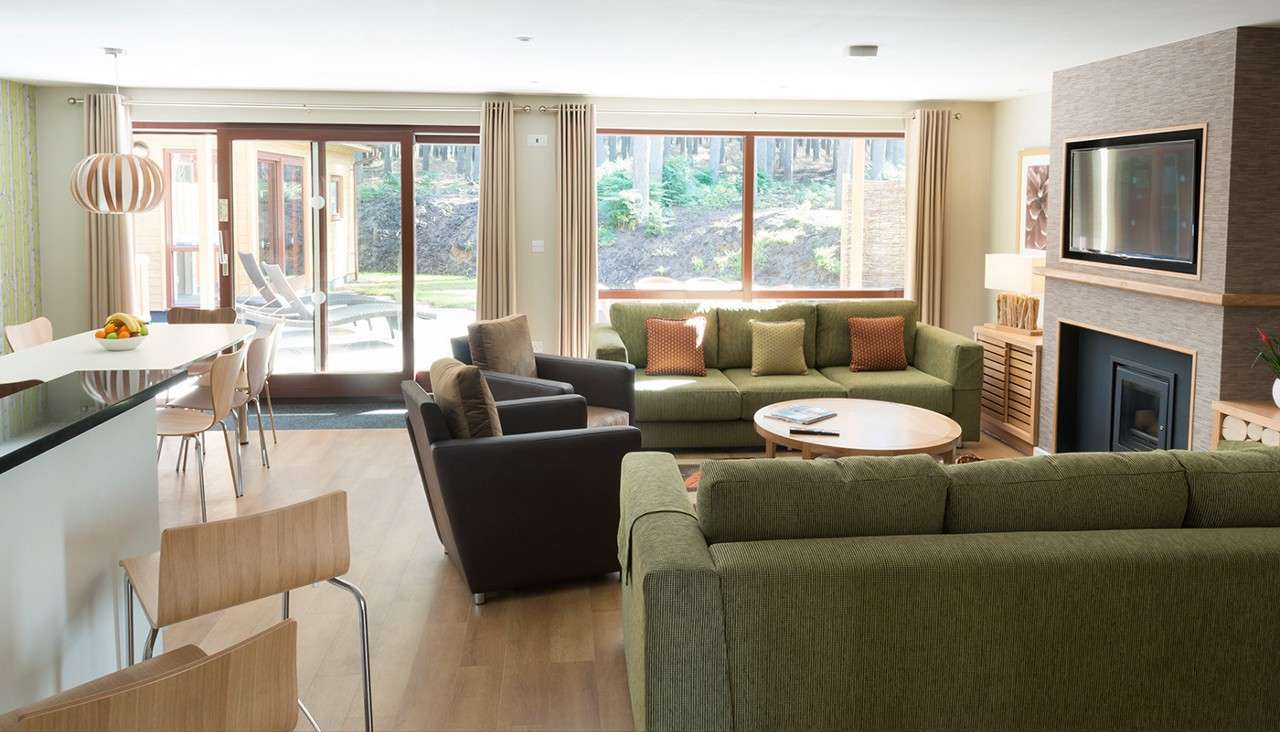 Green sectional sofa centers the space, facing a round coffee table and TV above a fireplace; armchairs, a kitchen island, and sliding glass doors lead to a patio and woods.
